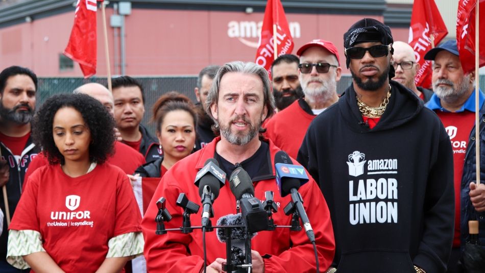 Gavin McGarrigle speaking into microphone with workers wearing red shirts and an Amazon facility in the background.
