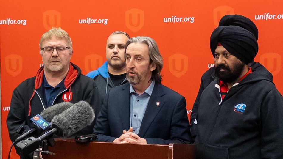 Gavin McGarrigle speaking into a mic at a podium surrounded by Unifor members and a red backdrop in behind.