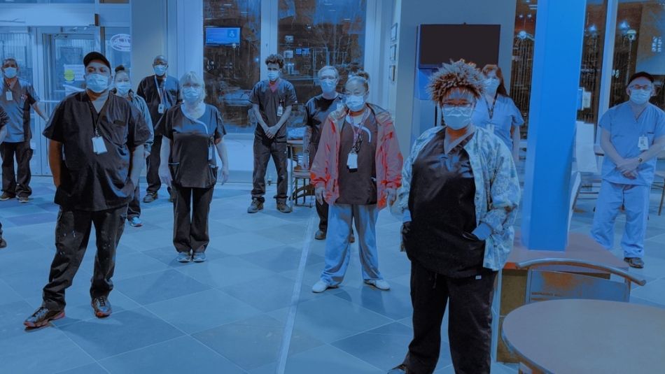 A group of healthcare workers in PPE stand socially distanced in the lobby of a healthcare facility.