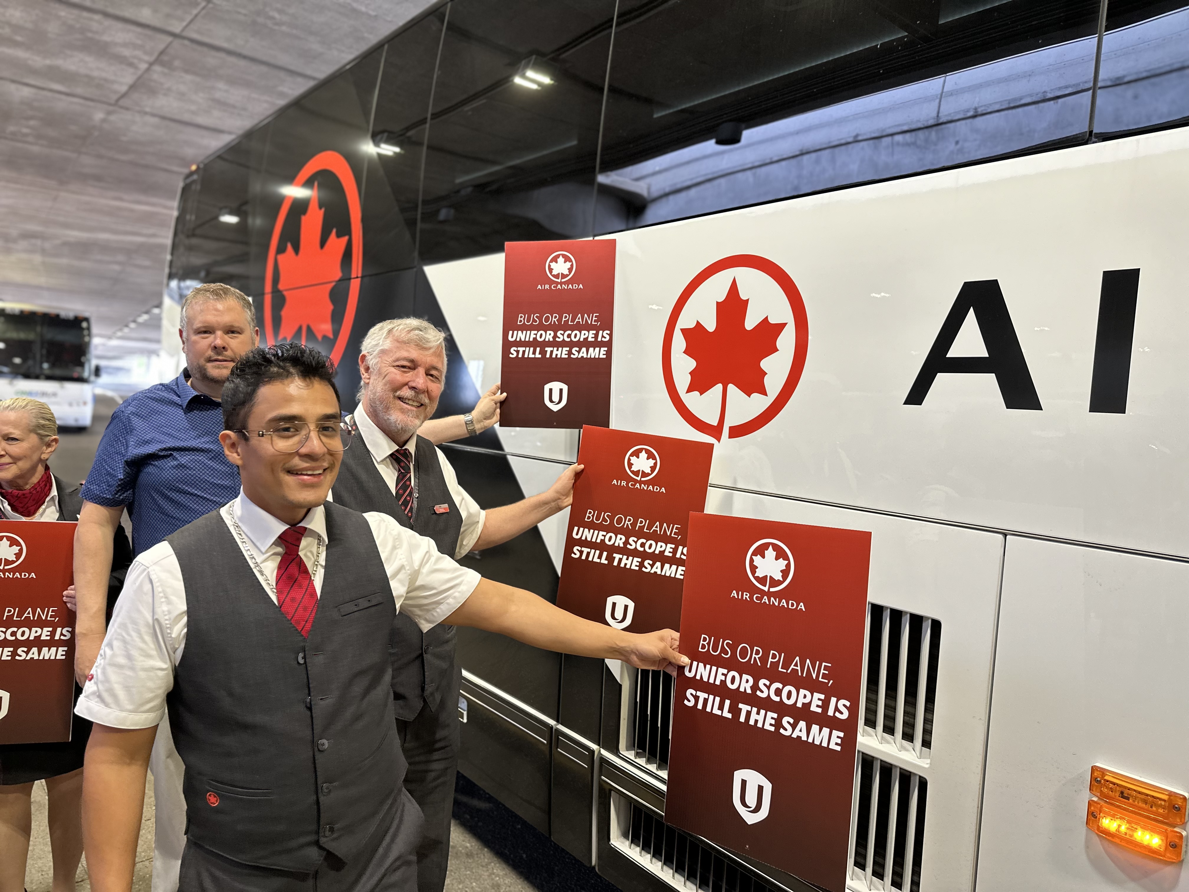Three men hold up place cards in front of a Air Canada bus that read : Bus or plane Unifor scope is the same