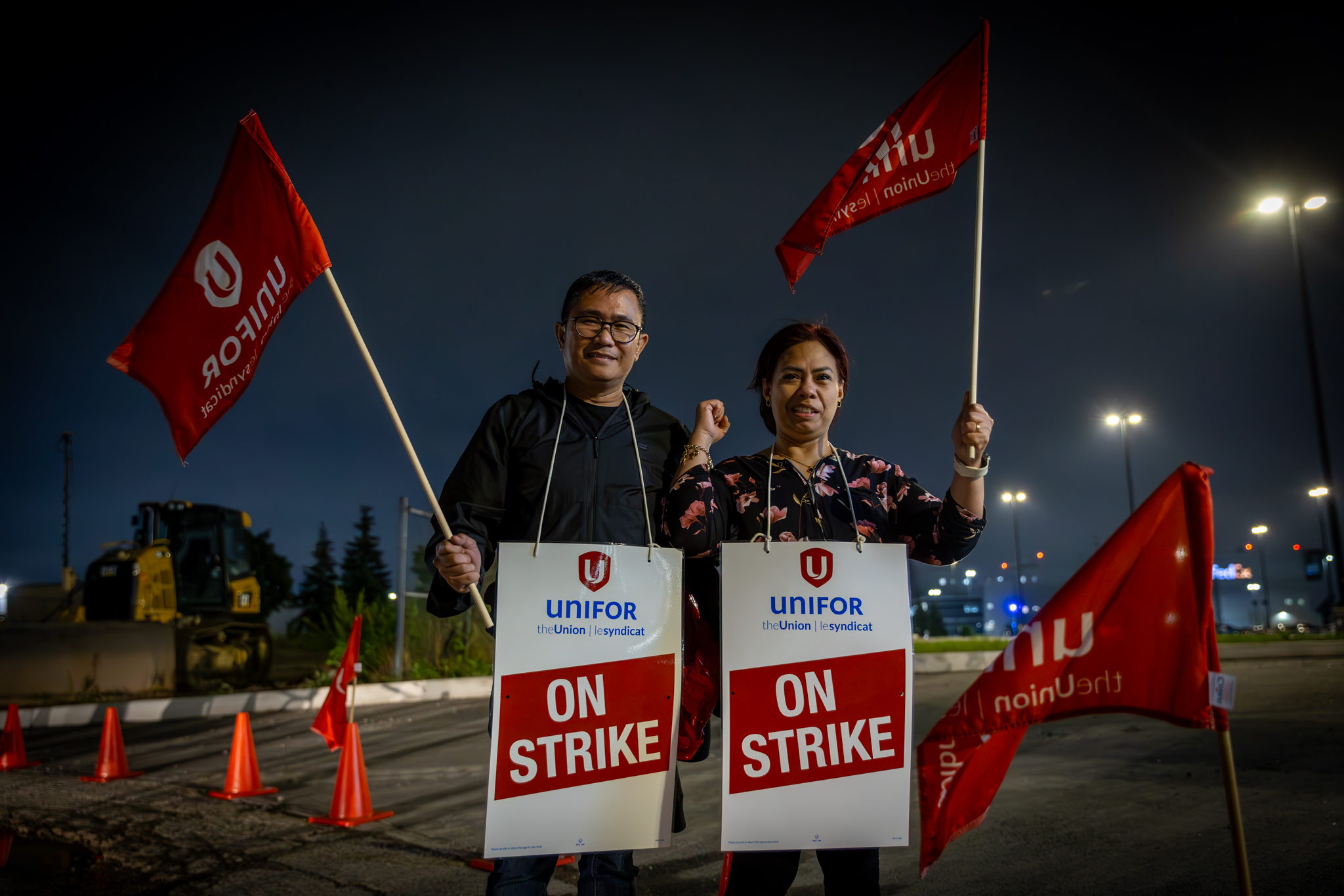 Two Unifor members standing on the Bombardier picket line wearing "on strike" signs and holding red Unifor flags.