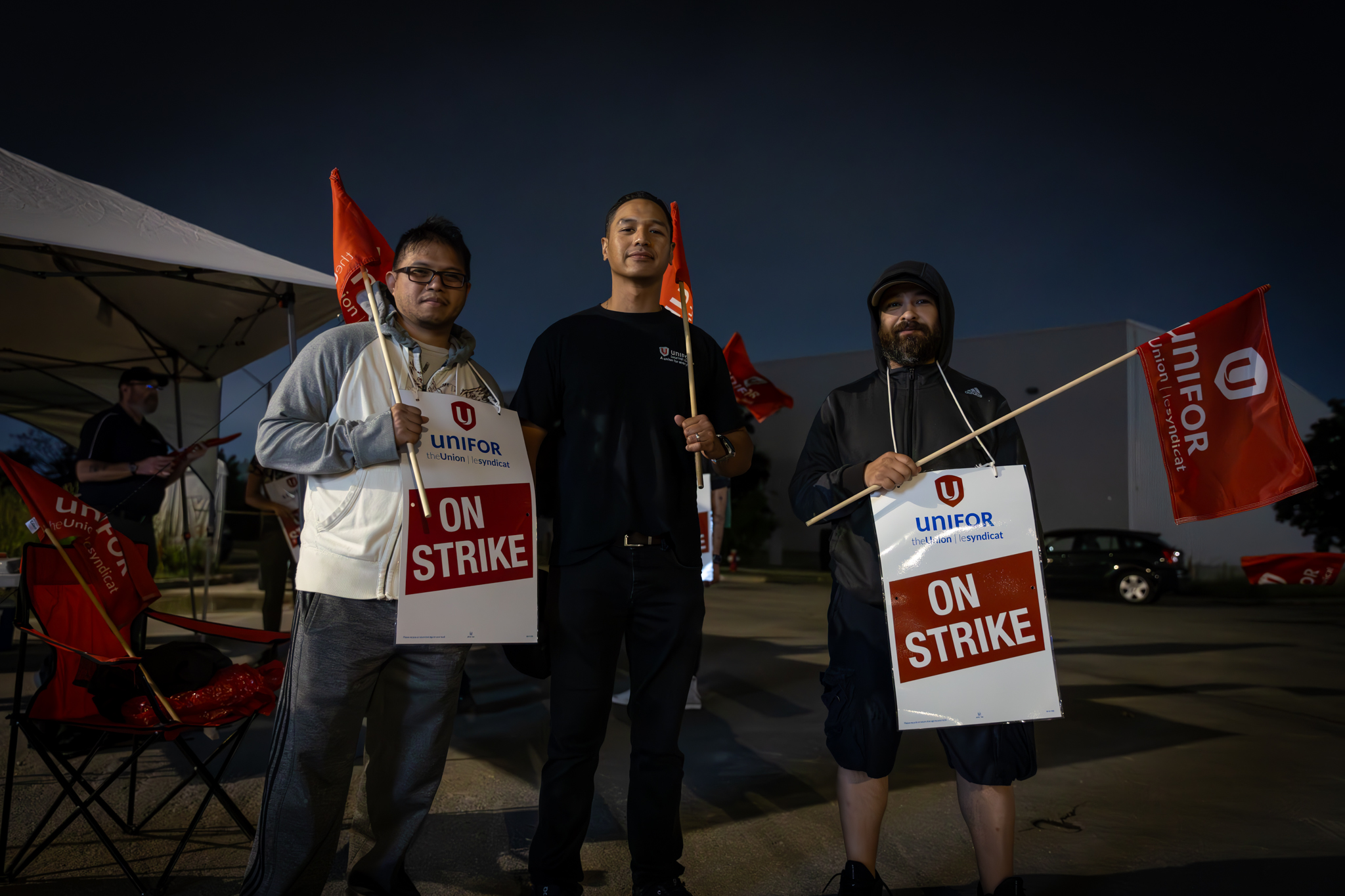 Three Unifor members wearing "on strike" signs and holding red Unifor flags standing on the Bombardier picket line.