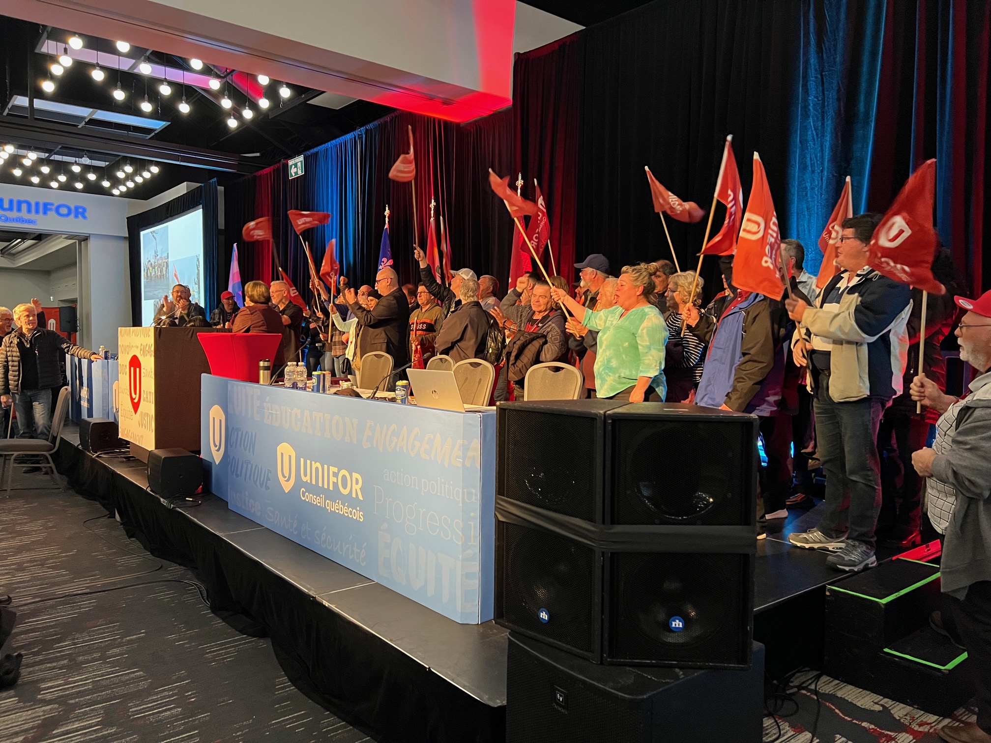 A group of people cheer and wave red Unifor flags while standing on a stage.