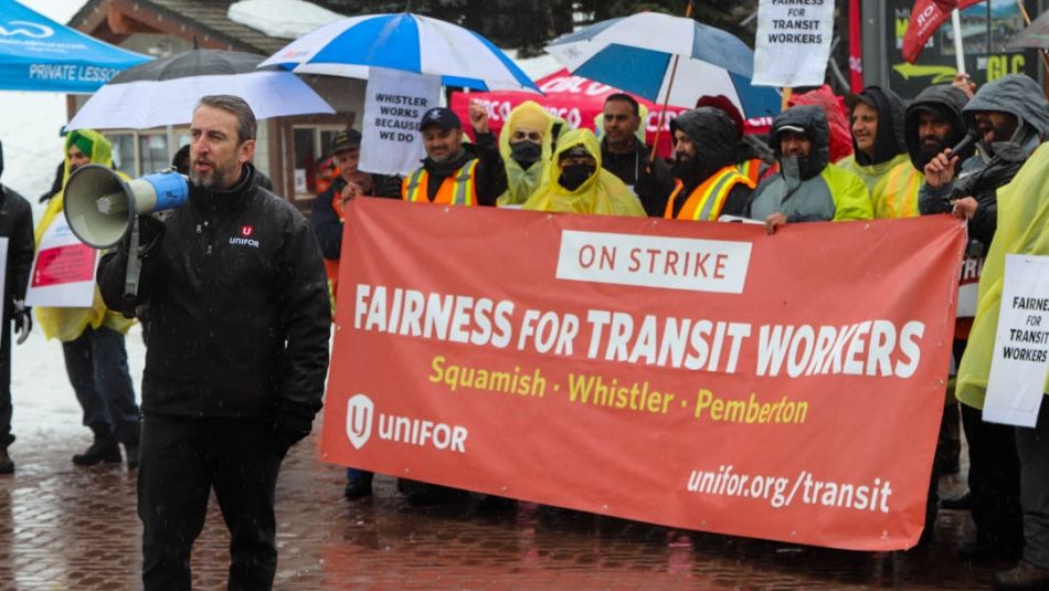 Un homme parle dans un porte-voix devant une foule et une bannière en soutien aux travailleuses et travailleurs du transport en commun qui demandent l’équité