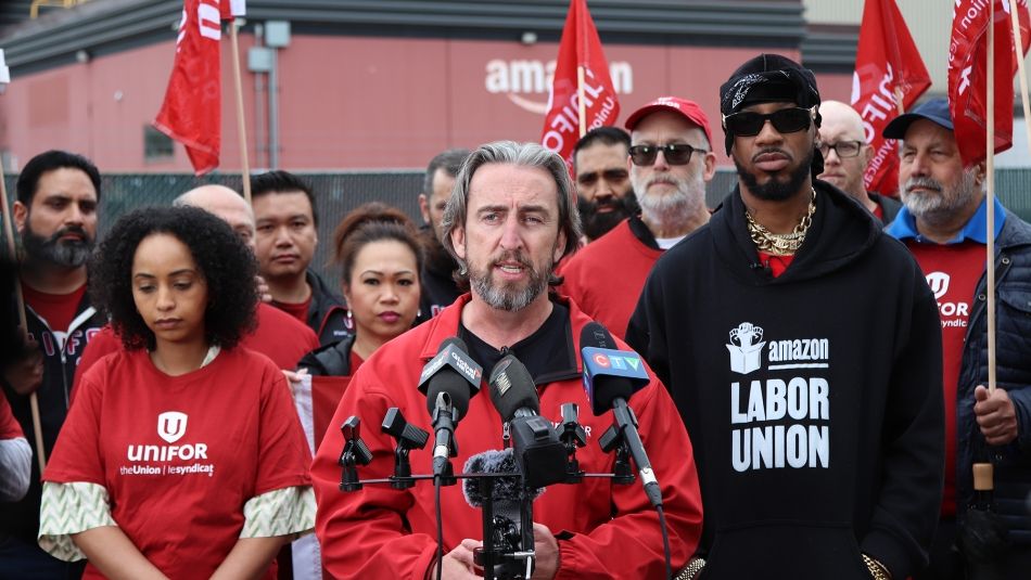 Gavin McGarrigle speaking at a mic with several workers standing behind him. An Amazon building is visible in the distance.