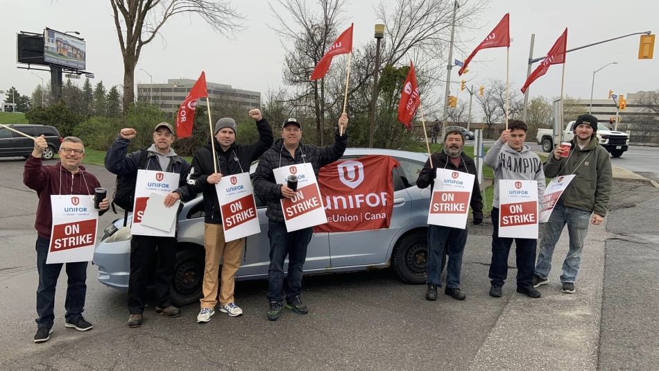 Seven people holding place cards that read "on strike" and red Unifor flags standing in front of a parked car