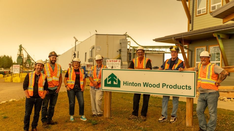 •	A group of people wearing safety vests and standing in front of a sign at Hinton Wood Products.