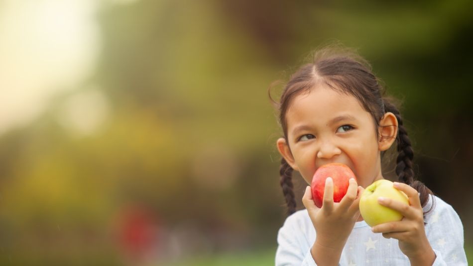 Girl with braids holding a yellow apple and biting into a red apple.