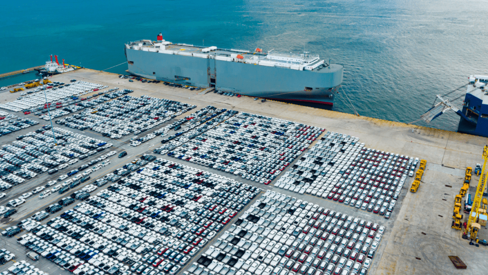 Overhead view of electric vehicles parked at a port awaiting to be loaded on to a card ship docked near by.