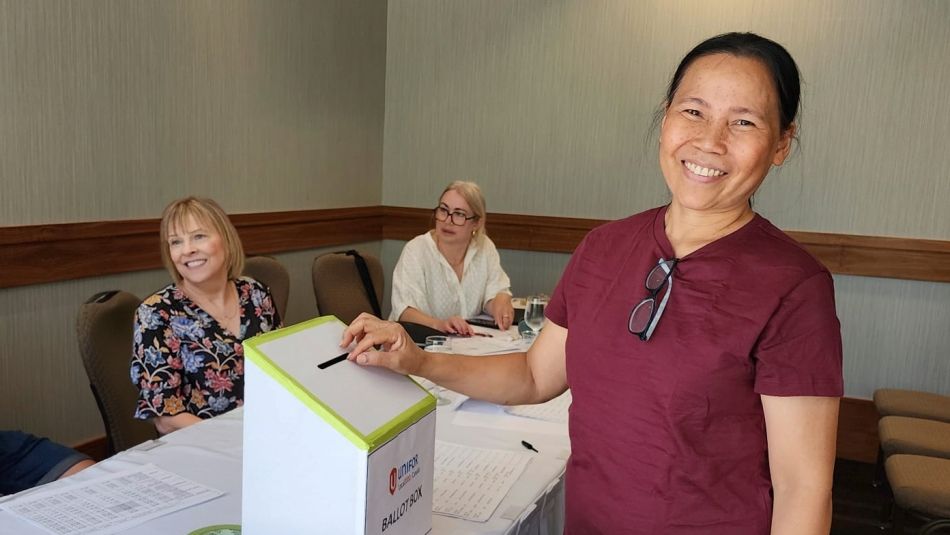 A smiling woman placing a ballot into a ballot box while others sit at a table nearby