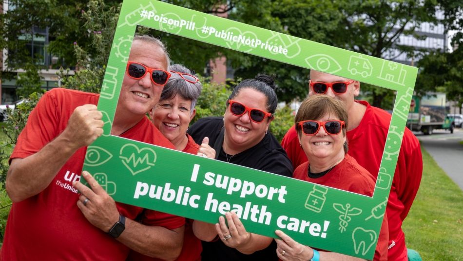 Five people in red shirts posing in a cutout selfie frame that reads: I support public health care