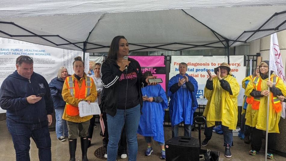 A women speaking at a mic under a tent. People standing in the back wearing rain coats