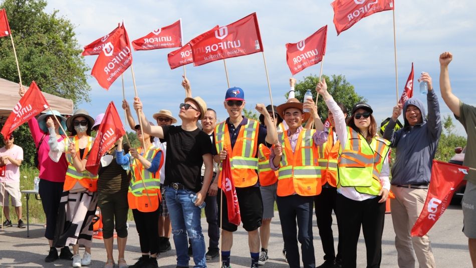 A line of Unifor members outdoors wearing high visibility vests and waving Unifor flags.