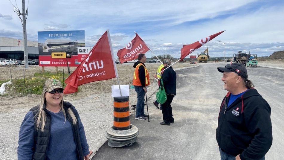 A group of people holding red union flags stand and converse on a road near construction equipment and a building, forming an picket line. One woman in a cap and sunglasses smiles while another man wears a black hoodie.