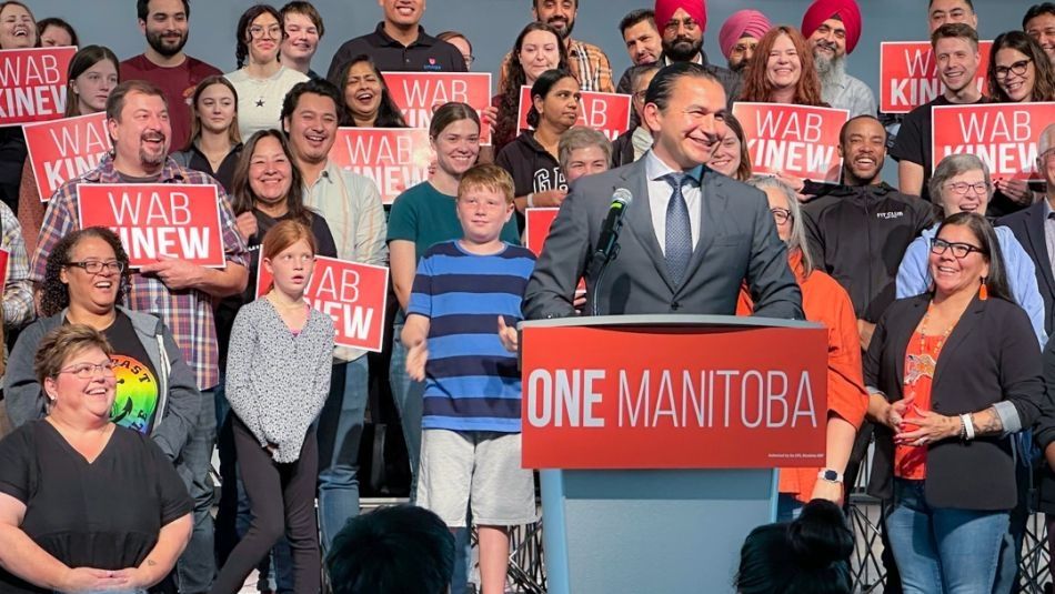 Wab Kinew speaks from a podium with a crowd of smiling supporters behind him, many holding 'Wab Kinew' signs.