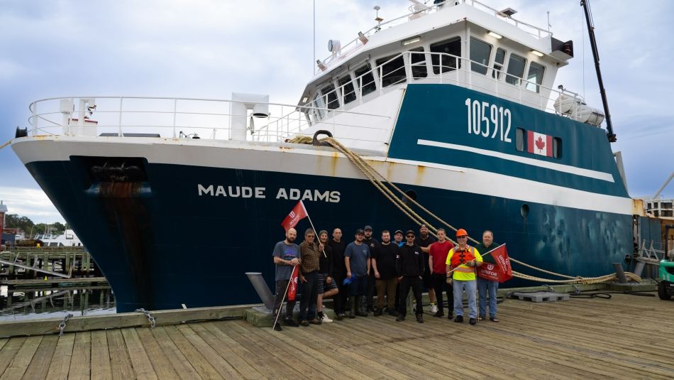 Ship crew stand on the wharf in front of the Maude Adams vessel, holding Unifor flags