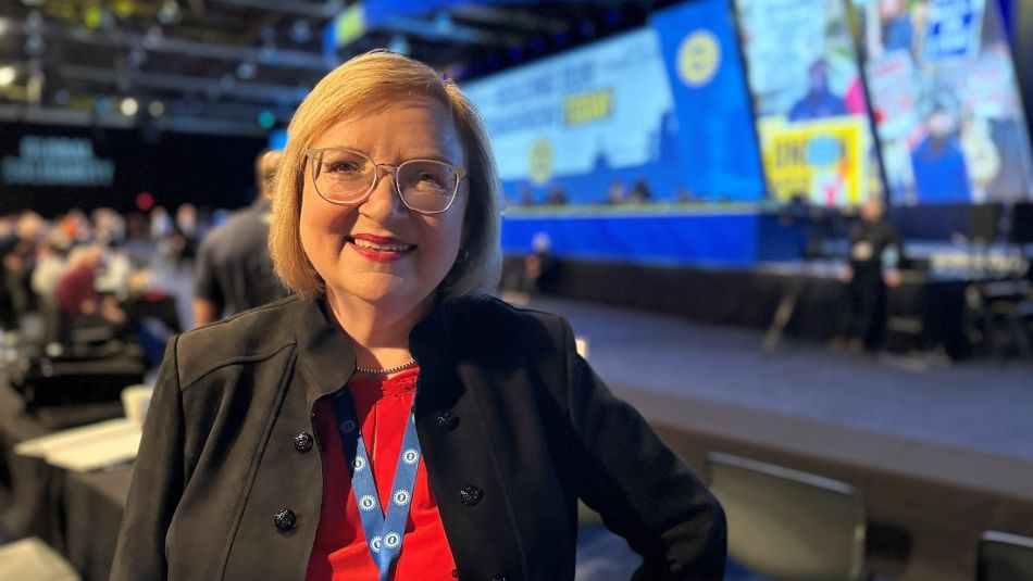 A woman wearing a red shirt and black blazer poses with screens and a panel table behind her in the background.