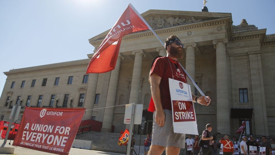 Beared man stands in front of the Manitoba legislature carrying a Unifor flag