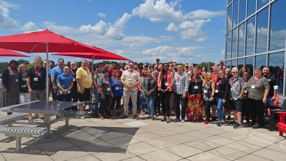A diverse group of people stands together on a rooftop terrace with red umbrellas, posing for a photo in front of a large glass building under a blue sky with scattered clouds.