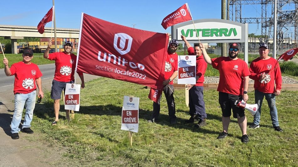 Six personnes portant des chemises rouges d'Unifor brandissent un grand drapeau rouge d'Unifor à l'extérieur, sur l'herbe, devant un bâtiment.