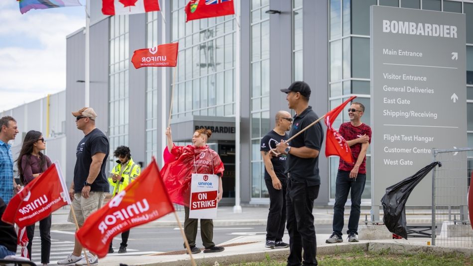 Protesters holding red Unifor flags stand outside a Bombardier building. One person waves a flag, while others hold signs with "ON STRIKE" messages. Canadian flags and a pride flag are visible. A large sign next to the building lists various entrances and facilities.
