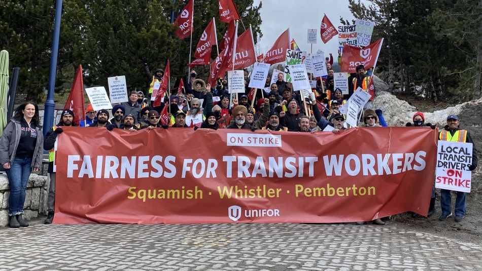 Large group of rally participants with flags and placards posing with a "Fairness for Transit Workers" banner on stairs outdoors.