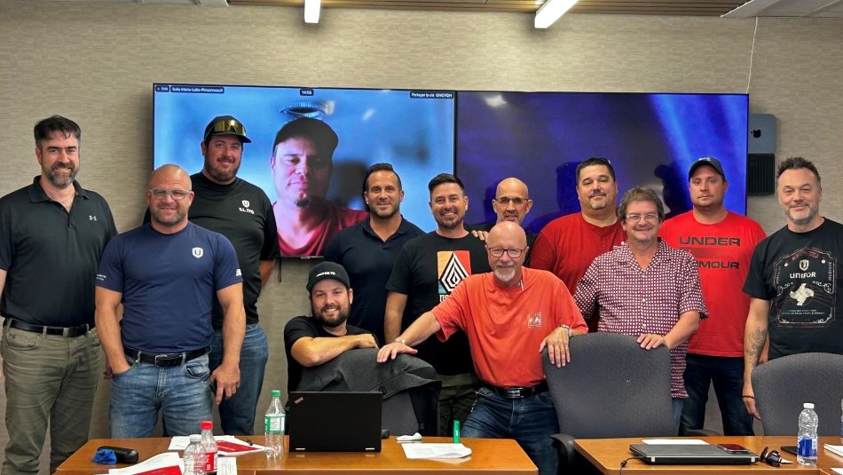 A large group of men standing behind desks posing for a photo in front of a TV where a man joins via Zoom.