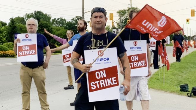 People standing on picket line with placards reading On Strike. Man in foreground is holding a Unifor flag