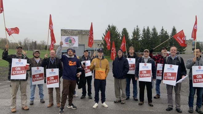 A group of workers stands outdoors on the picket line, holding "On Strike" signs and waving red flags. They appear to be part of a union protest or strike action. Trees and a building are visible in the background.