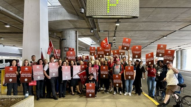 A large group of people standing in the airport under the P sign holding up Unifor flags and place cards that read "Bus or plane Unifor scope still the same"