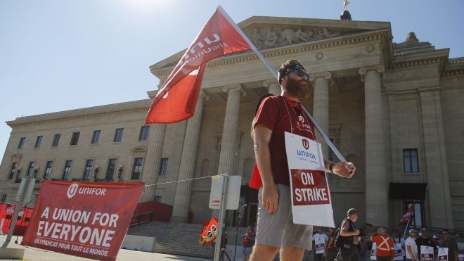 Beared man stands in front of the Manitoba legislature carrying a Unifor flag