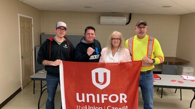 Four members of Unifor Local 31-X bargaining committee holding a large red Unifor banner. From left to right, a man in a baseball cap and hoodie, another in a blue t-shirt, a woman in a white blouse, and a man in a hi-vis vest and cap.