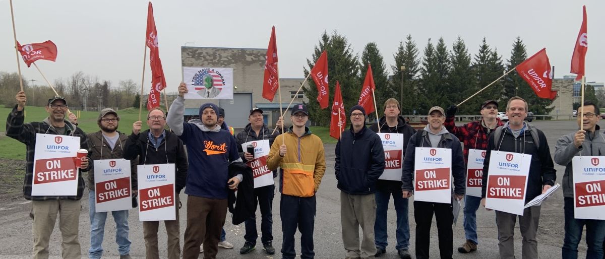 A group of people standing on a road holding red flags and signs that read "Unifor On Strike" forms a picketline. Trees and a building are visible in the background.